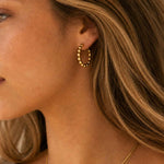 A close-up side profile of a woman wearing a small gold hoop earring with a textured beaded design and a delicate gold chain necklace.