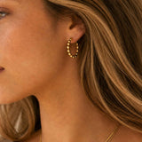 A close-up side profile of a woman wearing a small gold hoop earring with a textured beaded design and a delicate gold chain necklace.