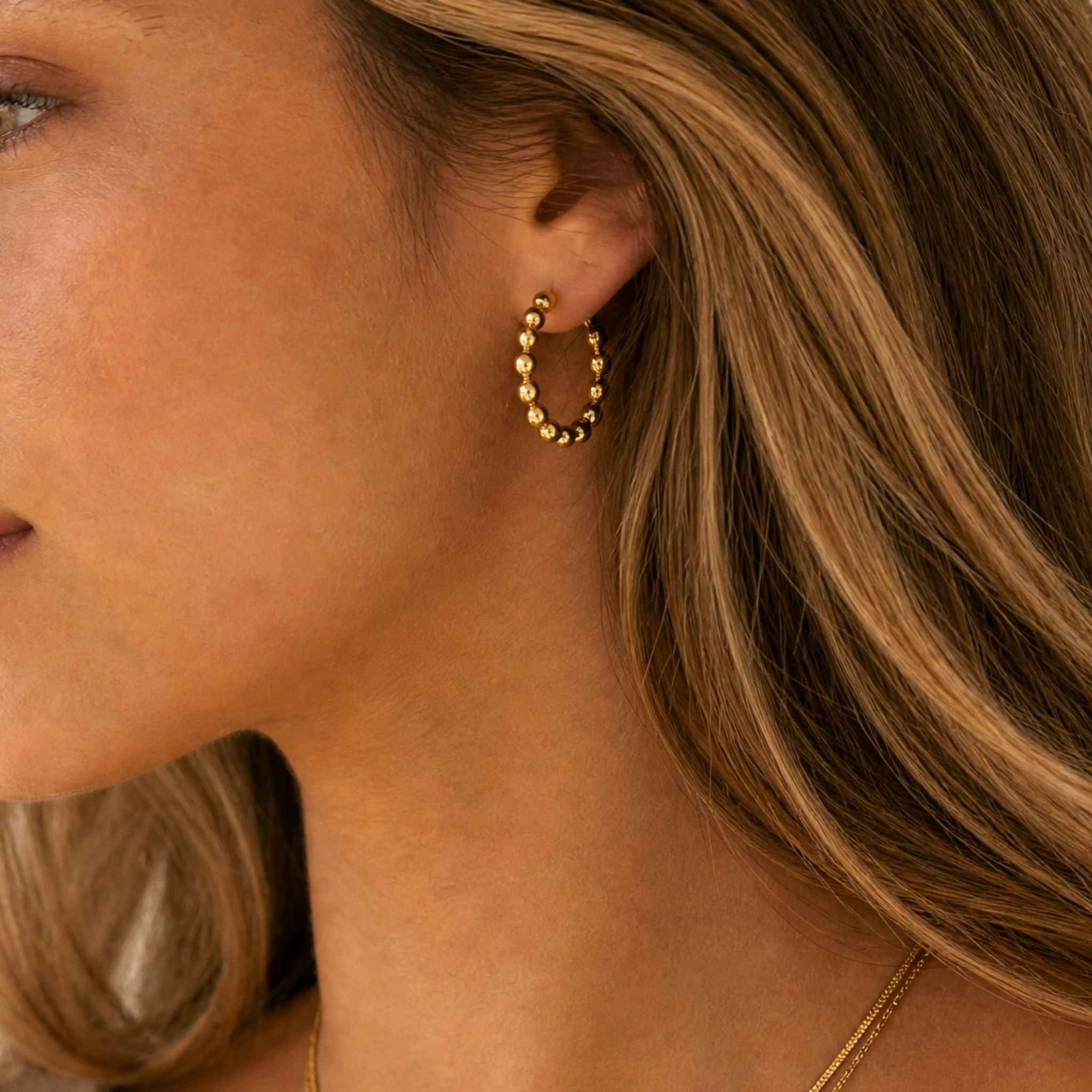 A close-up side profile of a woman wearing a small gold hoop earring with a textured beaded design and a delicate gold chain necklace.