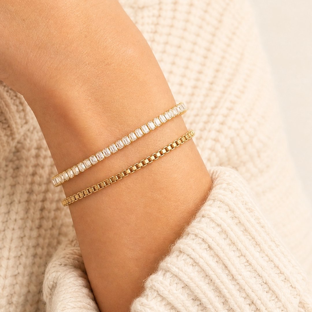 Two delicate bracelets worn on a wrist: one gold box chain bracelet and one gold tennis bracelet with clear stones, set against a cream knit sweater.