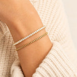 Two delicate bracelets worn on a wrist: one gold box chain bracelet and one gold tennis bracelet with clear stones, set against a cream knit sweater.