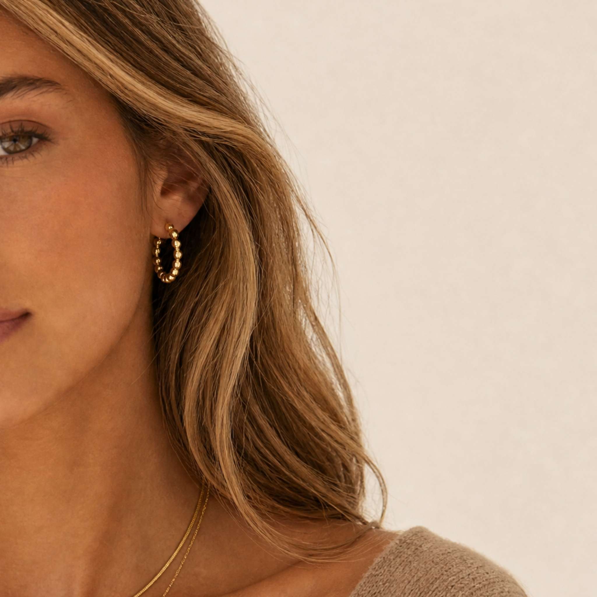 A close-up of a woman wearing a dainty gold hoop earring, with her face partially cropped on the left against a soft beige background.