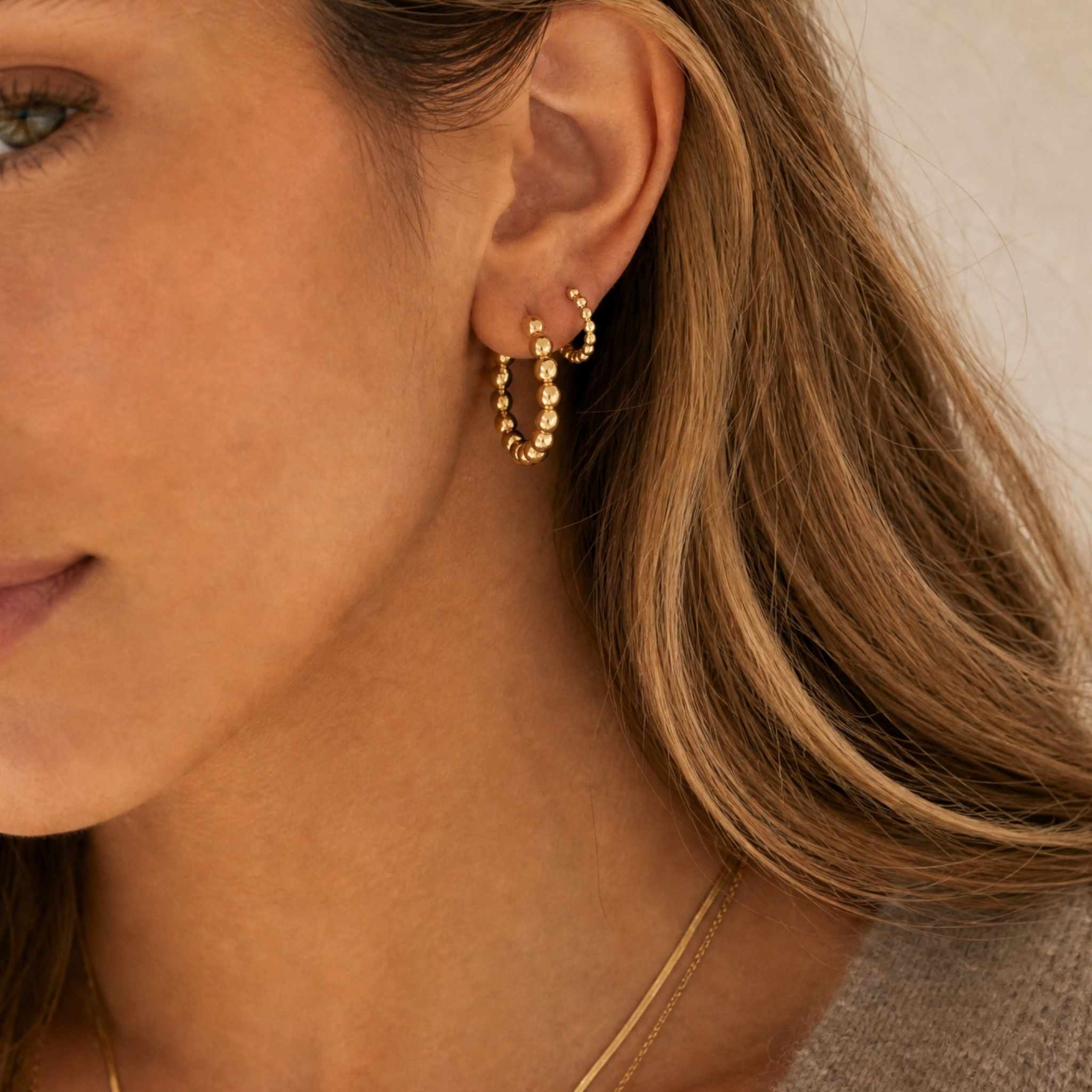 Close-up of a woman wearing small gold hoop earrings with a beaded texture, shown on her ear against her hair and skin.