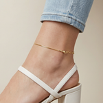 Gold anklet on a foot wearing a white sandal with a beige background
