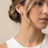 Close-up of a woman wearing a gold necklace and gold dainty earrings against a beige background