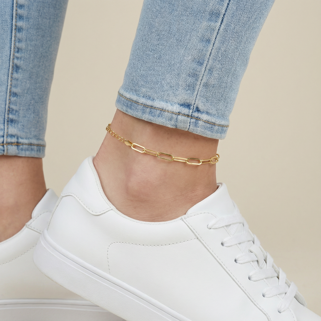 Gold paperclip anklet on a person's ankle with white sneakers and blue jeans.