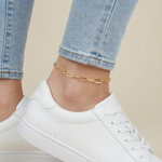 Gold paperclip anklet on a person's ankle with white sneakers and blue jeans.
