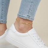 Gold paperclip anklet on a person's ankle with white sneakers and blue jeans.