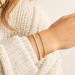 Two delicate gold bracelets worn on a woman's wrist, one with small clear stones and one with a textured box chain pattern, paired with a chunky cream knit sweater.