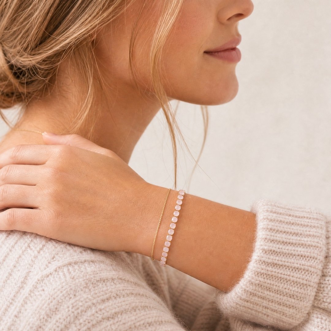 A close-up lifestyle shot of a woman wearing a delicate blush pink gold tennis bracelet on her wrist, paired with a cream knit sweater against a soft beige background.