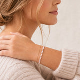 A close-up lifestyle shot of a woman wearing a delicate blush pink gold tennis bracelet on her wrist, paired with a cream knit sweater against a soft beige background.