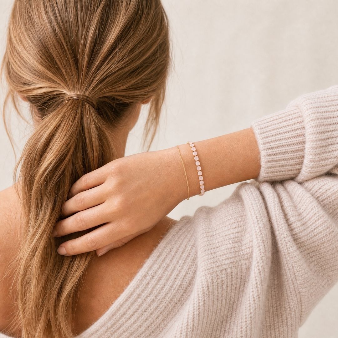 A woman shown from the back wearing a delicate pink gold tennis bracelet with small pink stones, paired with a matching thin gold chain. She is wearing a light beige knit sweater, and her hair is tied back in a low ponytail against a soft, light beige background.