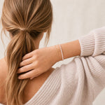 A woman shown from the back wearing a delicate pink gold tennis bracelet with small pink stones, paired with a matching thin gold chain. She is wearing a light beige knit sweater, and her hair is tied back in a low ponytail against a soft, light beige background.
