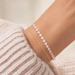 A close-up of a person's wrist wearing two delicate bracelets: one pink stone tennis bracelet and one thin gold chain, styled with a ribbed cream knit sweater.