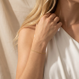 Close-up of a person wearing a dainty gold bracelet on a neutral background
