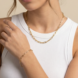 Gold chain necklace and paperclip bracelet on a person wearing a white top against a beige background