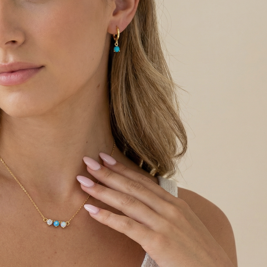 Close-up of a woman wearing gold turquoise huggie earrings and an opal turquoise necklace on a beige background