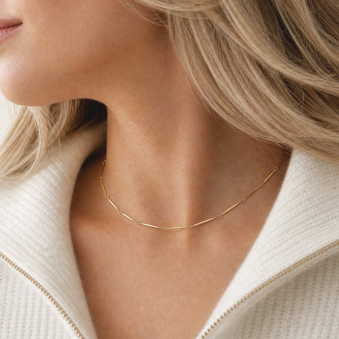 A close-up of a woman's neck and collarbone wearing a tarnish free gold chain necklace with small spaced links, paired with a cream-colored ribbed knit top.