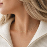 A close-up of a woman's neck and collarbone wearing a tarnish free gold chain necklace with small spaced links, paired with a cream-colored ribbed knit top.
